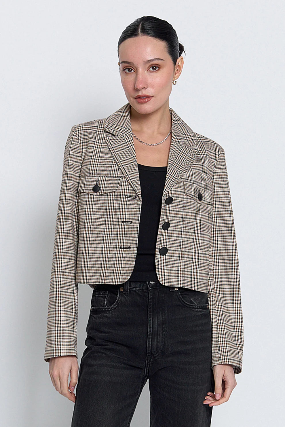 A woman modeling a cropped, long-sleeve plaid blazer in beige, brown, and black, worn over a black top and dark jeans against a light gray studio background.