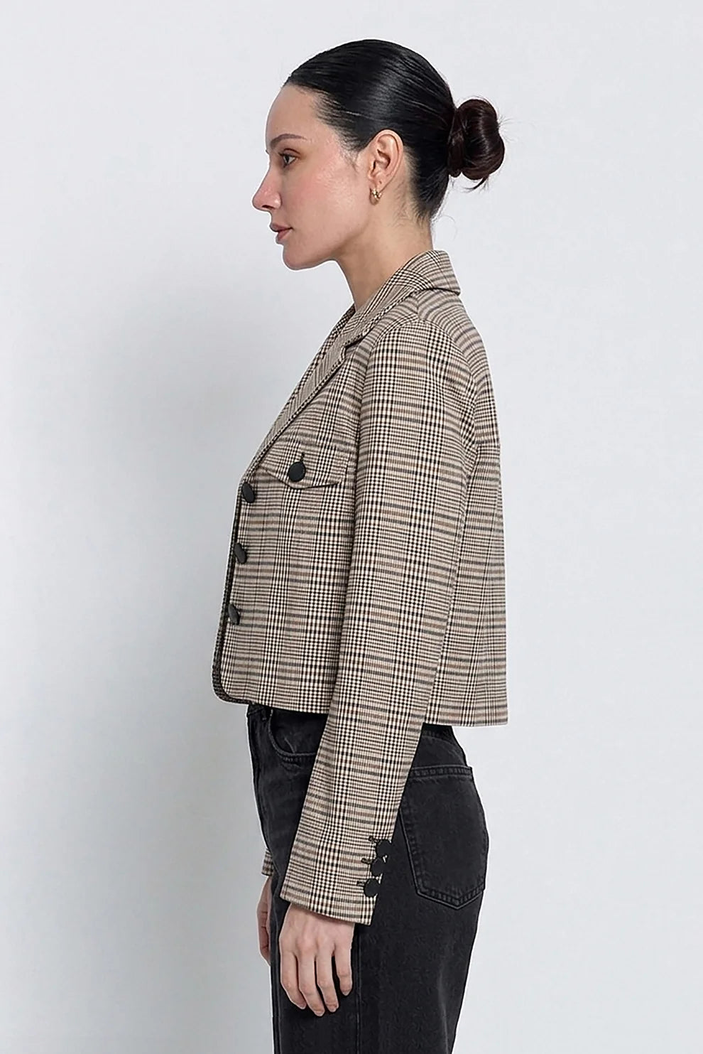 A woman in profile wearing a cropped, double-breasted plaid blazer in beige and brown tones, paired with black jeans, against a plain light gray background.