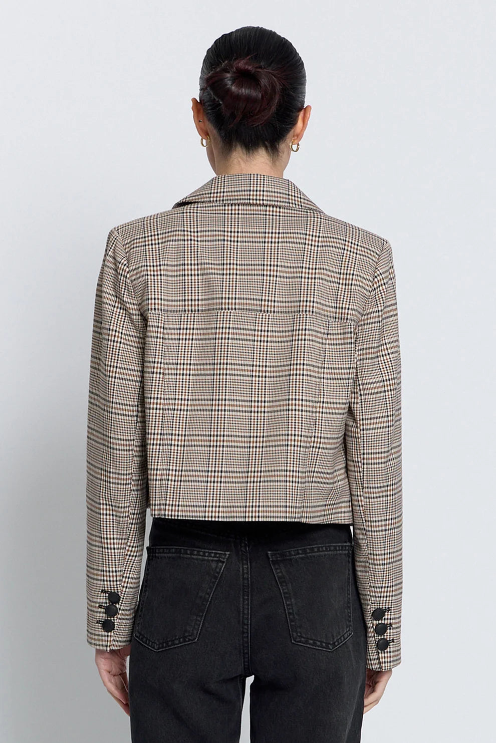 A woman in back view wearing a cropped, double-breasted plaid blazer in beige and brown tones, paired with black jeans, against a plain light gray background.