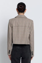 A woman in back view wearing a cropped, double-breasted plaid blazer in beige and brown tones, paired with black jeans, against a plain light gray background.
