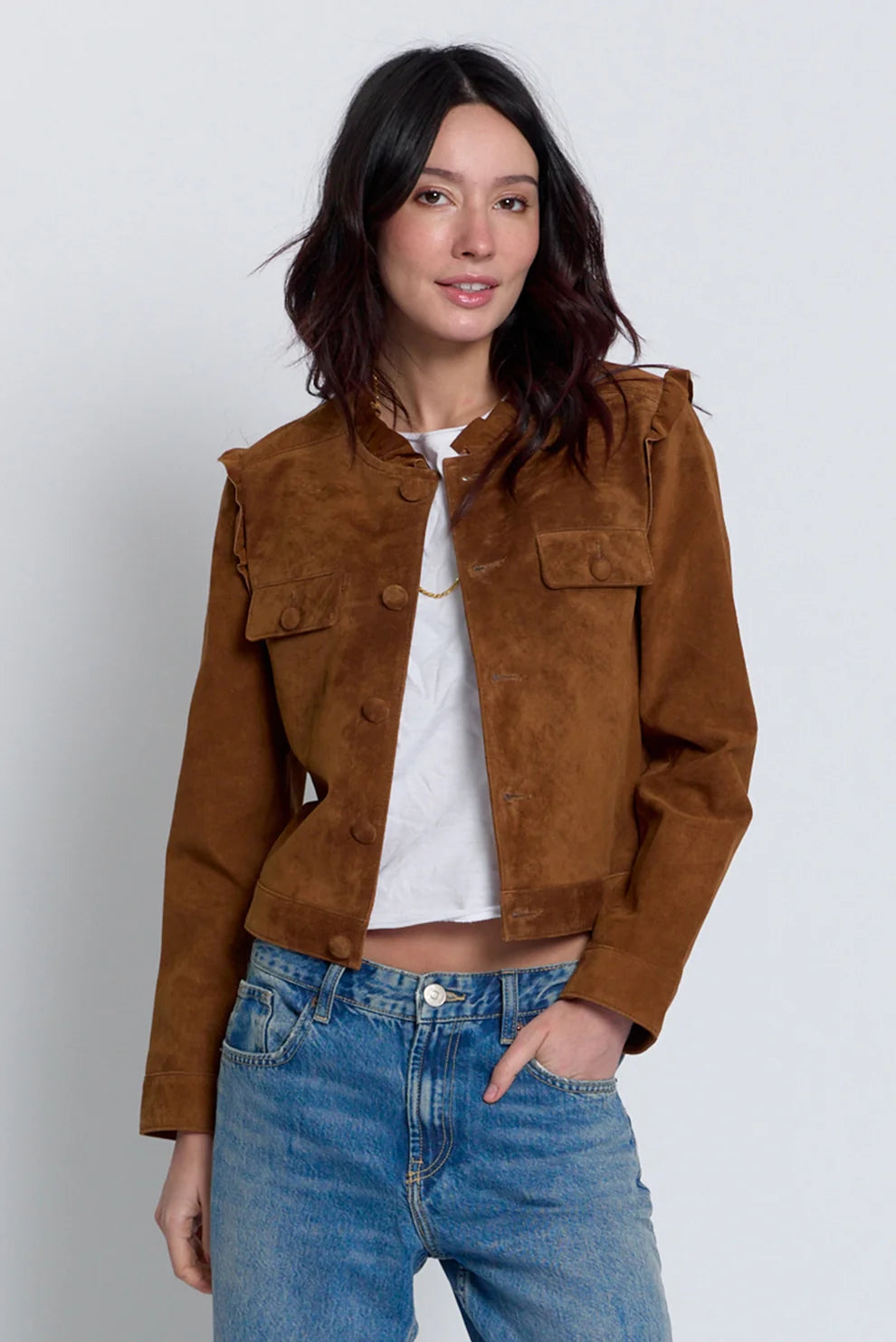 A woman modeling a cropped brown suede jacket with ruffle details on the shoulders, worn over a white crop top and blue jeans, against a light gray studio background.