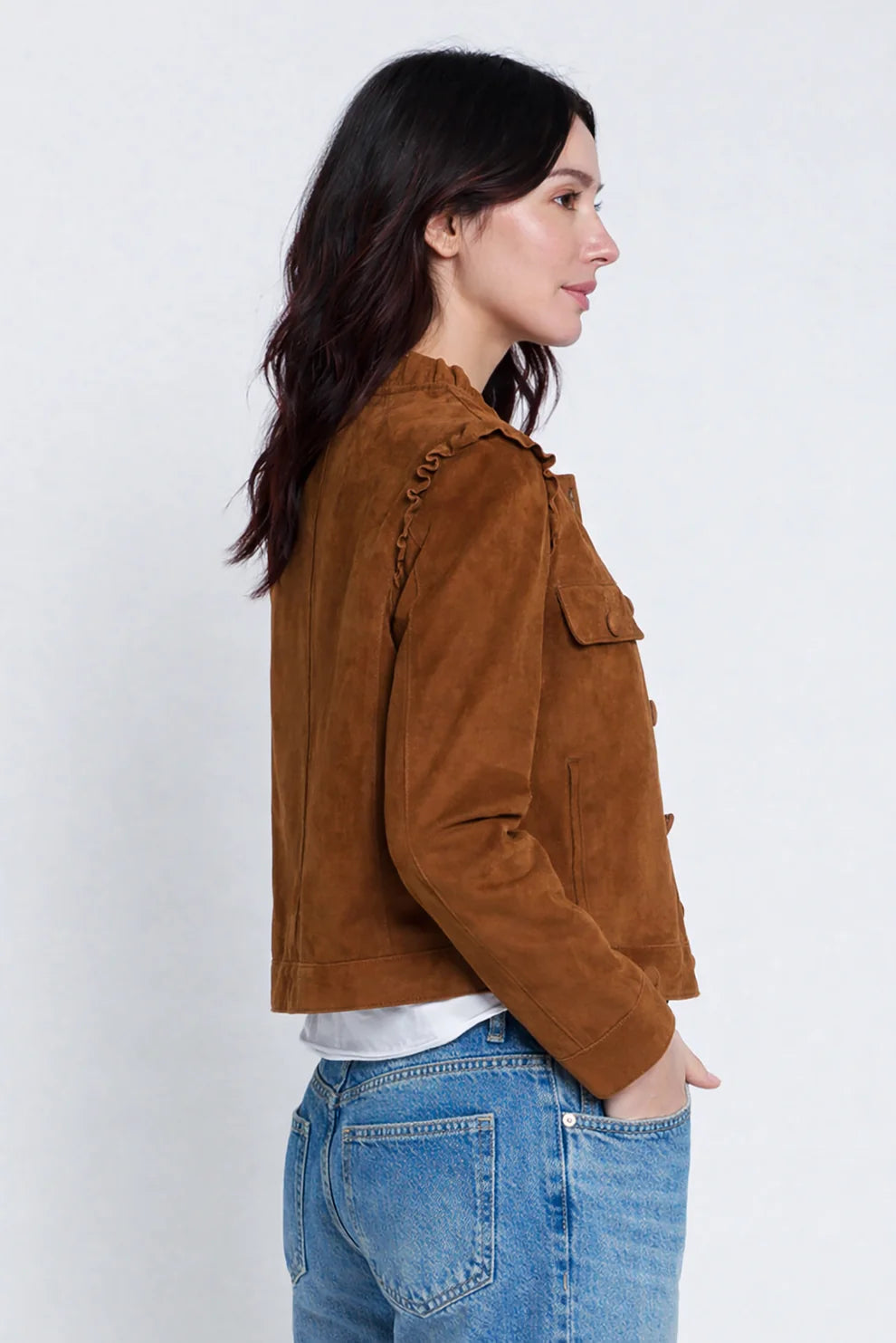 A woman shown in side profile wearing a cropped brown suede jacket over a white top and blue jeans, photographed against a plain light gray studio background.