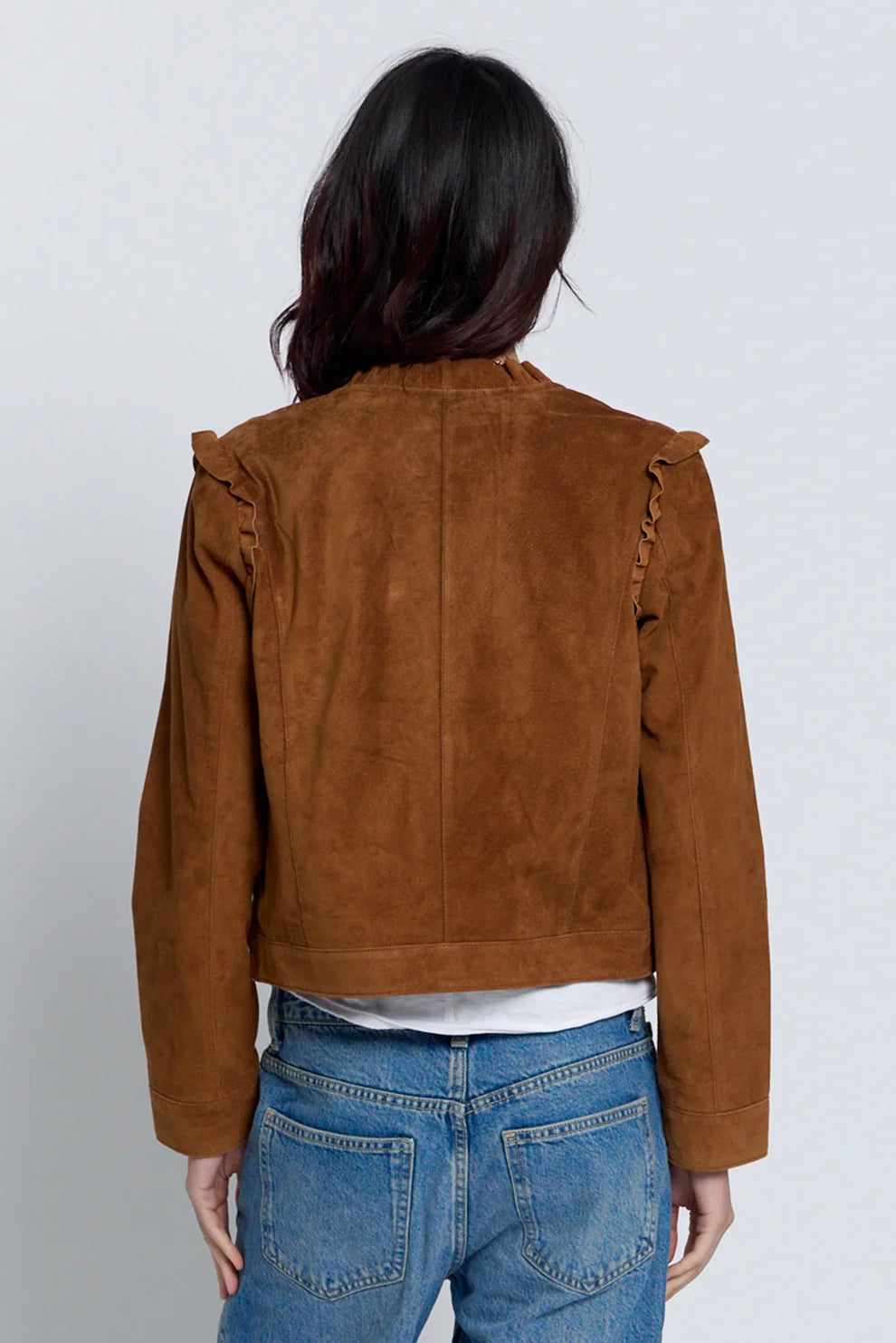 A woman viewed from the back wearing a cropped brown suede jacket with decorative fringed details on the shoulders, paired with blue jeans and a white t-shirt, against a light gray studio background.
