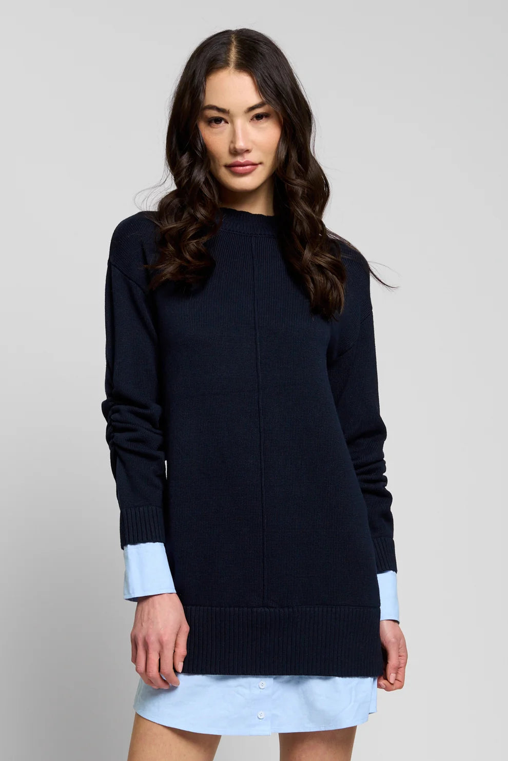 A woman modeling a dark navy blue knit sweater dress with light blue shirt cuffs peeking out at the sleeves, worn over a light blue shirt and skirt, against a solid light gray background.