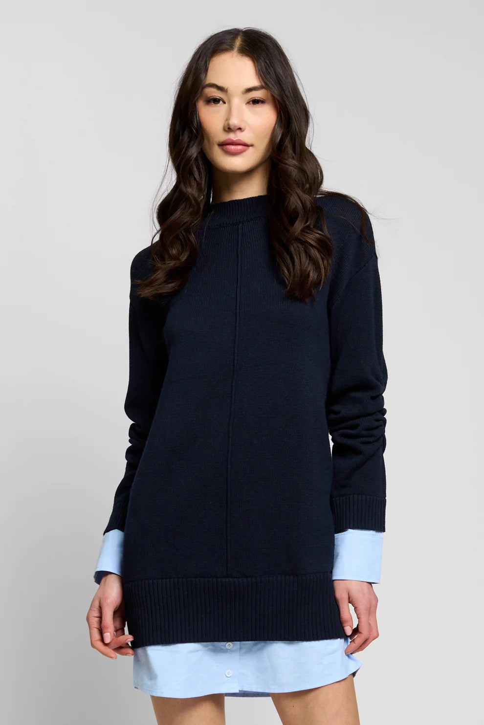 A woman modeling a dark navy blue mock-neck knit sweater with light blue contrast cuffs, layered over a light blue shirt dress, against a plain light gray studio background.