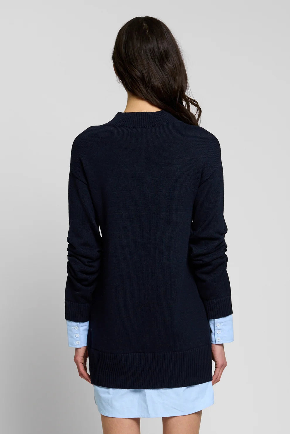 A woman viewed from the back wearing a dark navy knit sweater over a light blue shirt, against a plain light gray studio background.