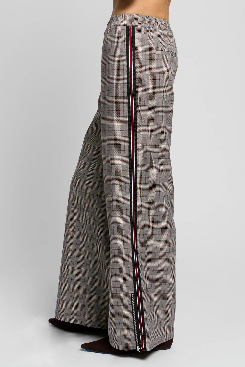 A side view of a person wearing wide-leg plaid trousers in gray, beige, and brown, featuring a vertical striped accent down the side.