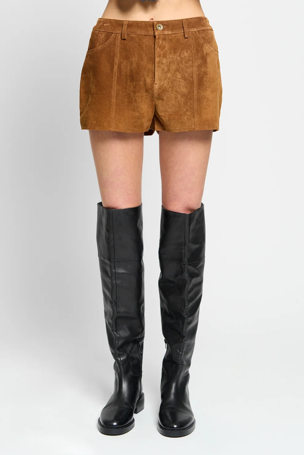 A model from the waist down wearing brown suede shorts and black leather over-the-knee boots, photographed against a light gray studio background.