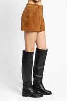 A model from the waist down wearing brown suede shorts and black knee-high leather boots, photographed against a clean white studio background.