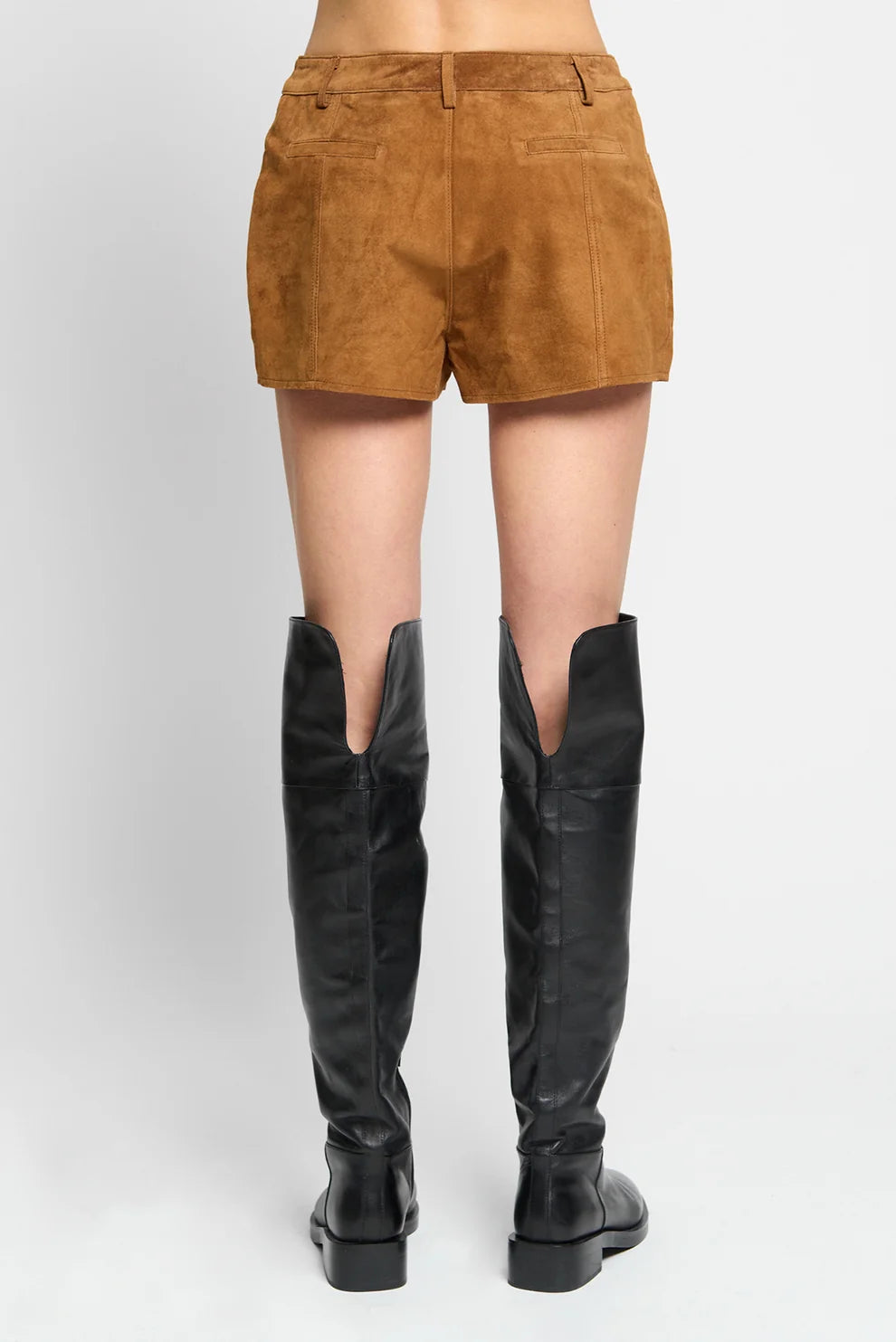 A person shown from the back wearing tan suede shorts and black over-the-knee boots against a light gray studio background.