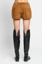 A person shown from the back wearing tan suede shorts and black over-the-knee boots against a light gray studio background.