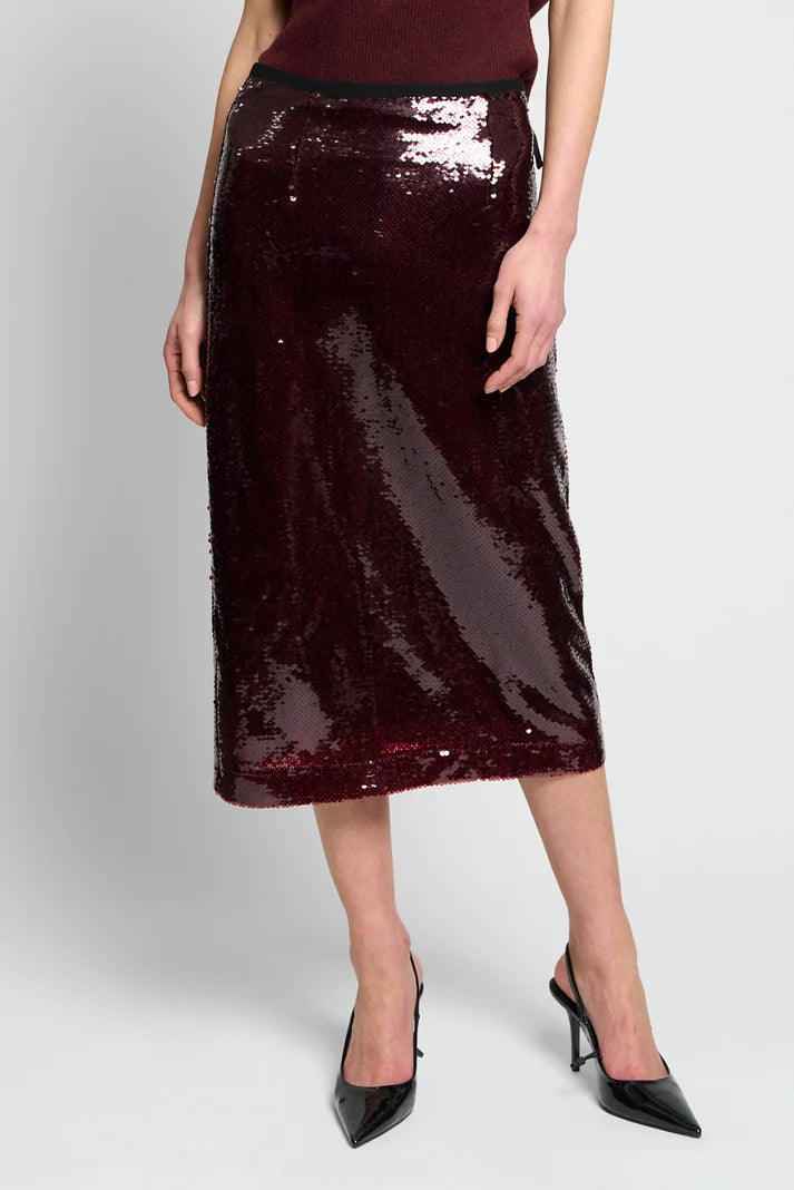 A model wearing a dark burgundy sequined midi skirt with a black waistband, paired with black pointed-toe heels, against a light gray studio background.