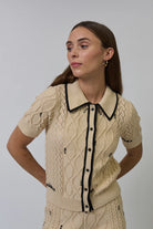 Woman wearing a beige cable knit cardigan with black trim on a plain background