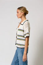 Woman wearing a white crochet top with rainbow stripes and blue jeans on a light gray background