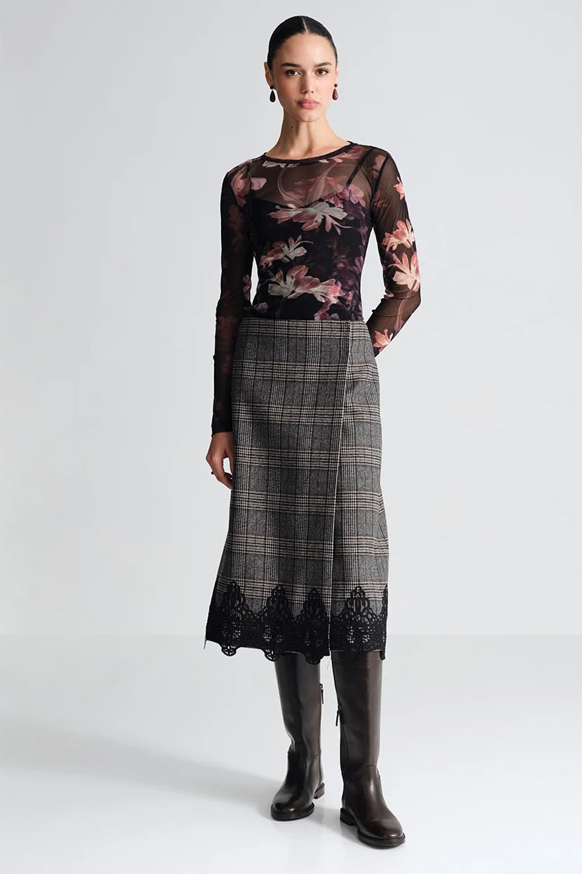 A model stands against a light gray studio background wearing a sheer long-sleeve floral top with a gray plaid skirt featuring black lace trim, paired with dark knee-high boots.