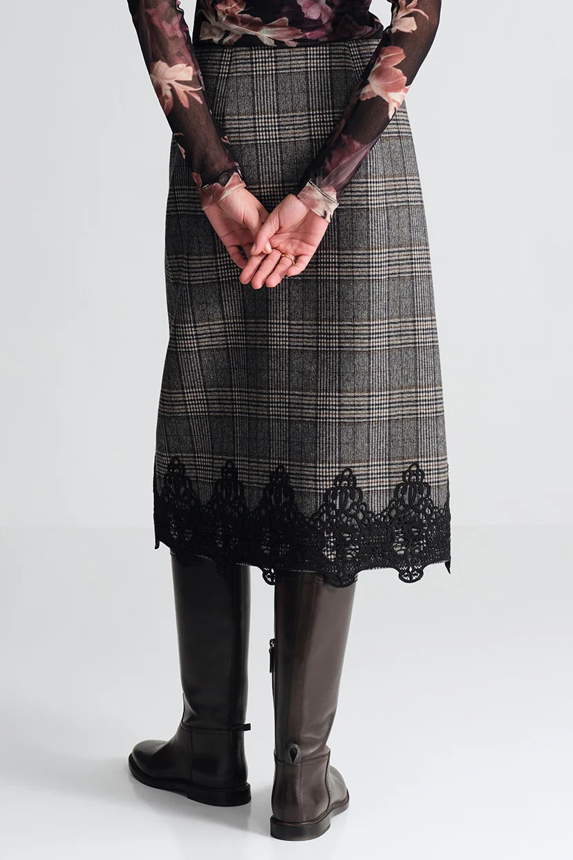 A person shown from the back wearing a knee-length plaid skirt with a black lace hem, paired with dark brown knee-high boots and a floral long-sleeve top, against a light gray studio background.