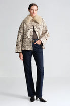 A model stands against a light gray studio background wearing a beige and brown patterned jacket with a large cream fur collar, dark blue jeans, and black pointed shoes.