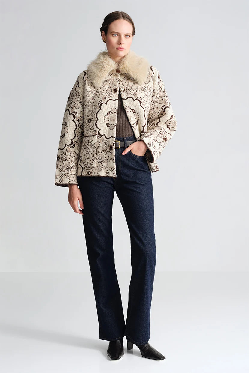 A model stands against a light gray studio background wearing a beige and brown patterned jacket with a large cream fur collar, dark blue jeans, and black pointed shoes.