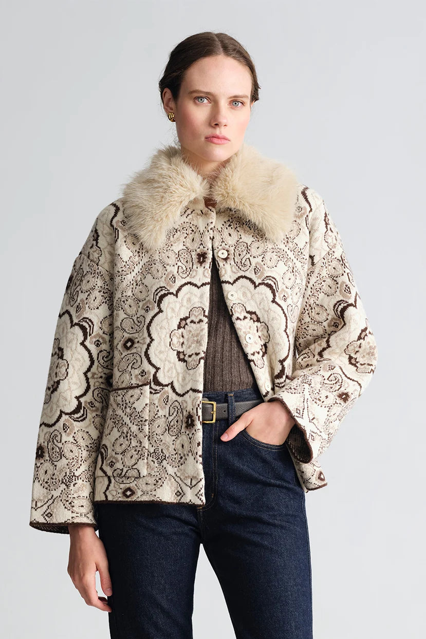 A model wearing a cropped beige quilted jacket with a brown paisley pattern, a large cream faux-fur collar, and dark blue jeans, photographed against a neutral light gray background.