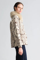 A woman shown in side profile wearing a beige and brown patterned jacket with a fluffy cream fur collar, paired with dark blue jeans, against a light gray studio background.