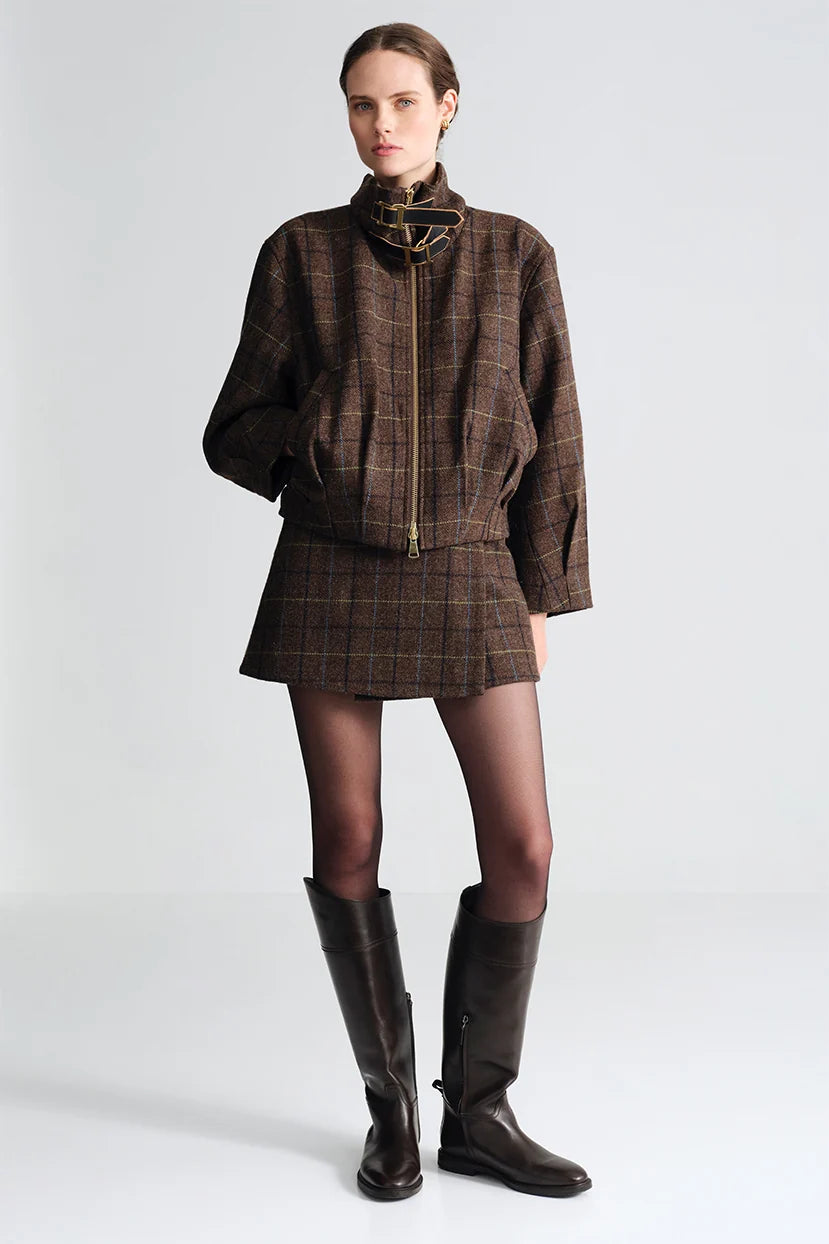 A model stands against a light gray studio background wearing a brown plaid jacket and matching mini skirt set, paired with sheer black tights and dark brown knee-high boots.