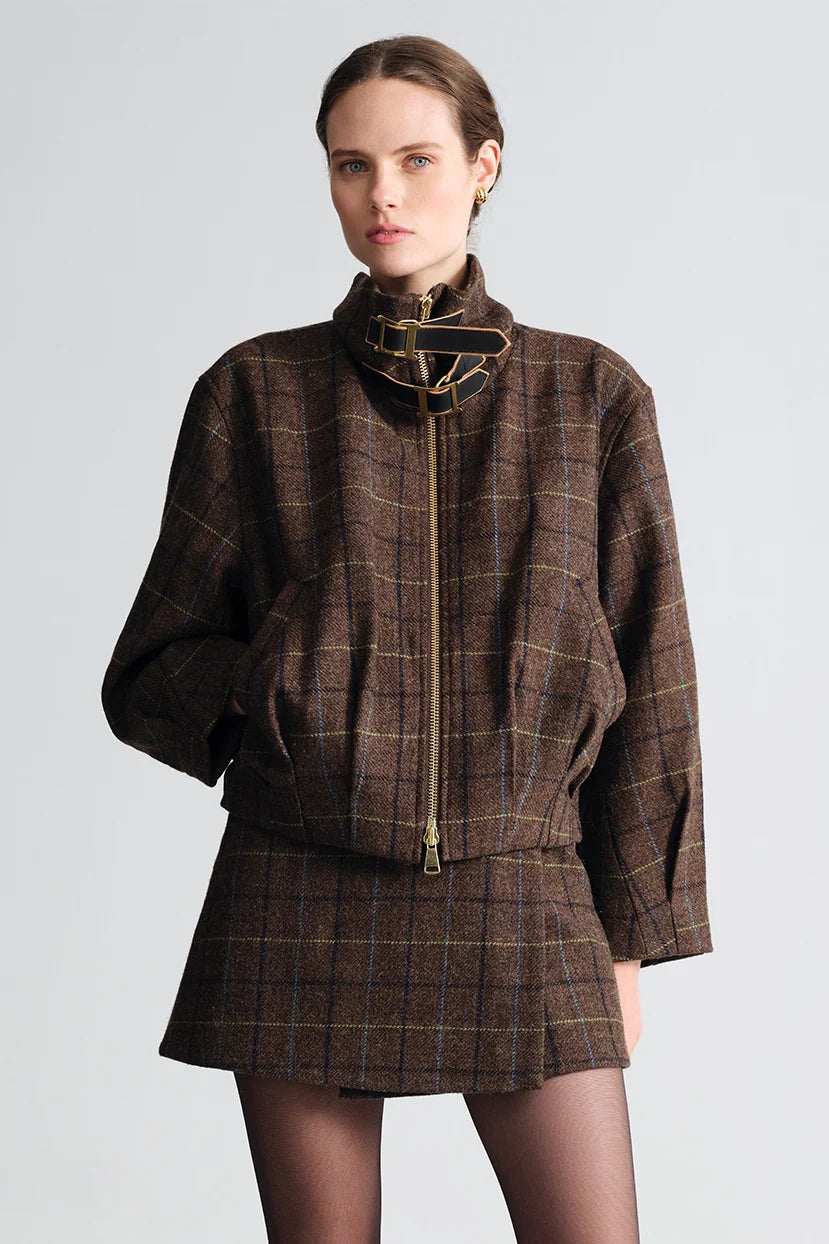 A model wearing a brown plaid jacket and matching mini skirt set with a high mock neck and gold-tone zipper details, photographed against a light gray studio background.