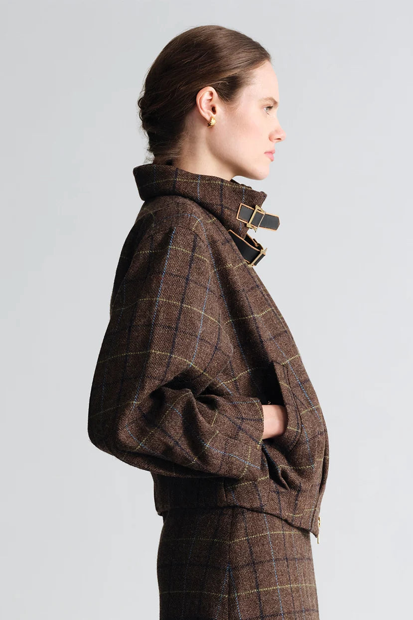 A woman shown in side profile wearing a brown plaid cropped jacket with a high structured collar and matching trousers, photographed against a light gray studio background.