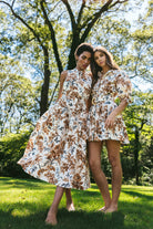 Two women in floral dresses standing in a park with trees and grass in the background.