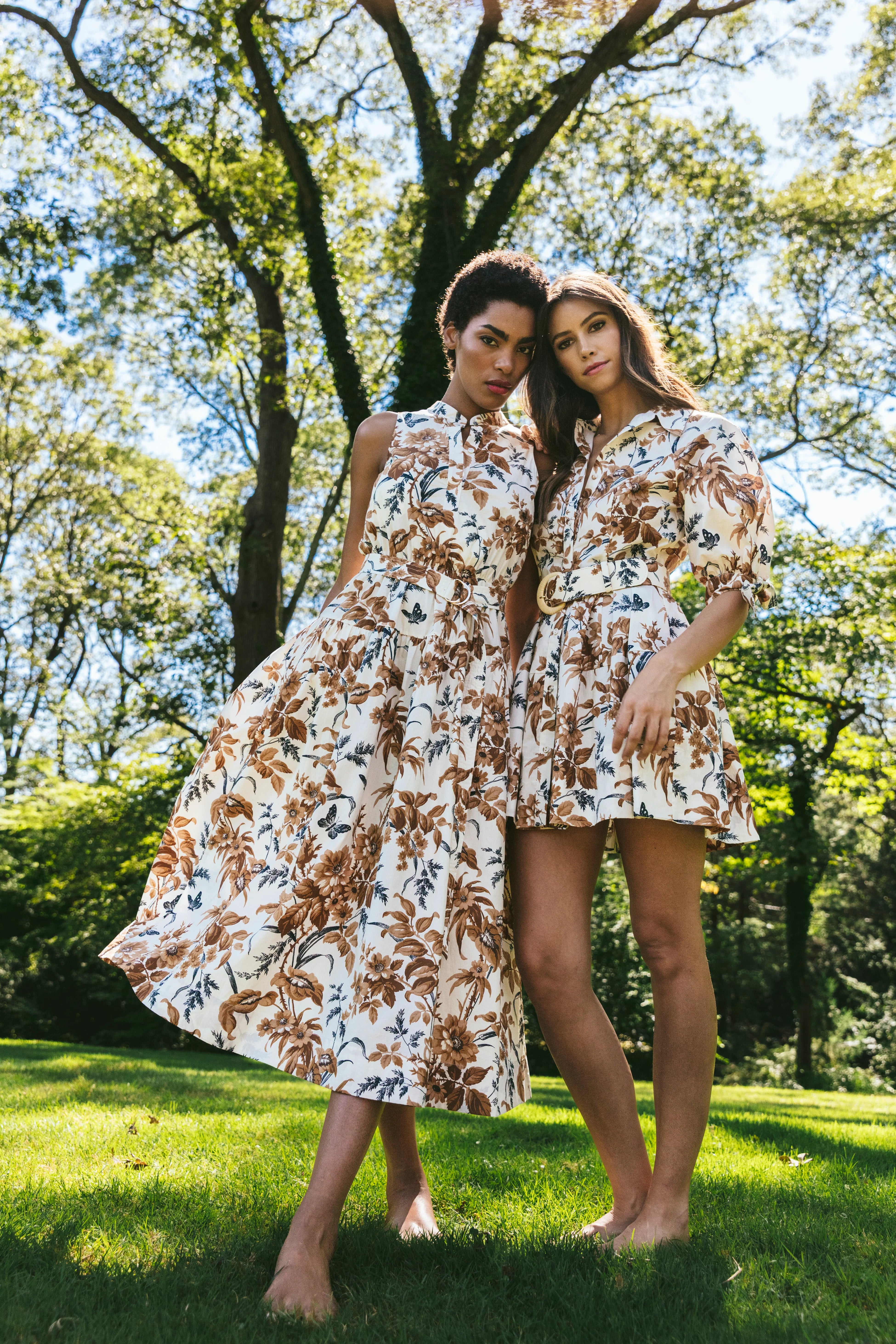 Two women in floral dresses standing in a park with trees and grass in the background.
