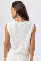 Woman wearing a white sleeveless top with intricate detailing on a plain background