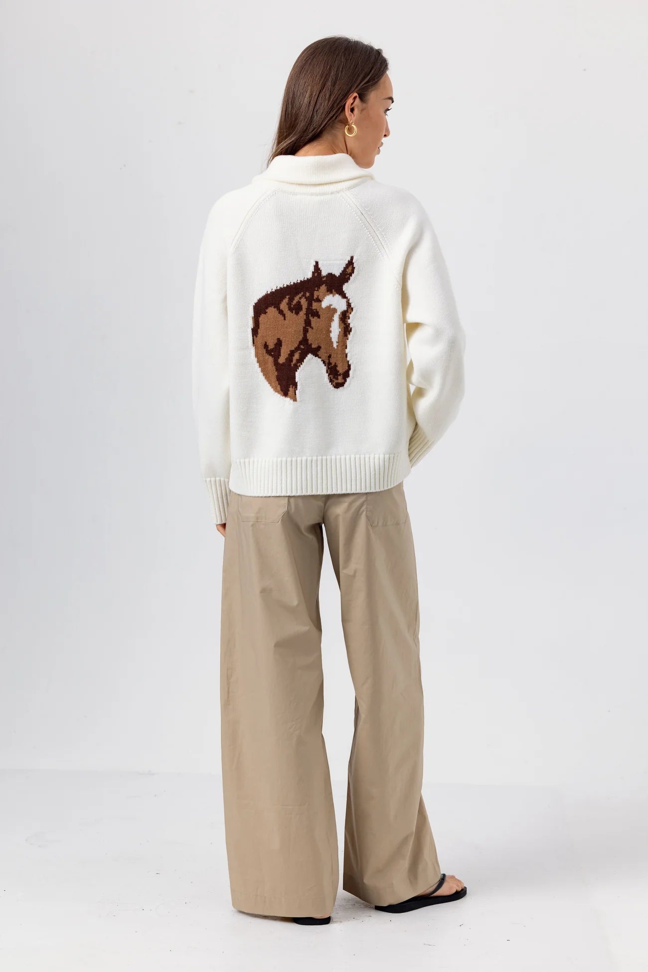 Woman wearing a white sweater with a horse design and beige pants on a white background