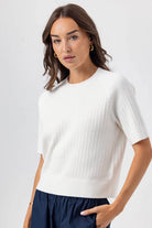 Woman wearing a white sweater and navy pants against a plain background