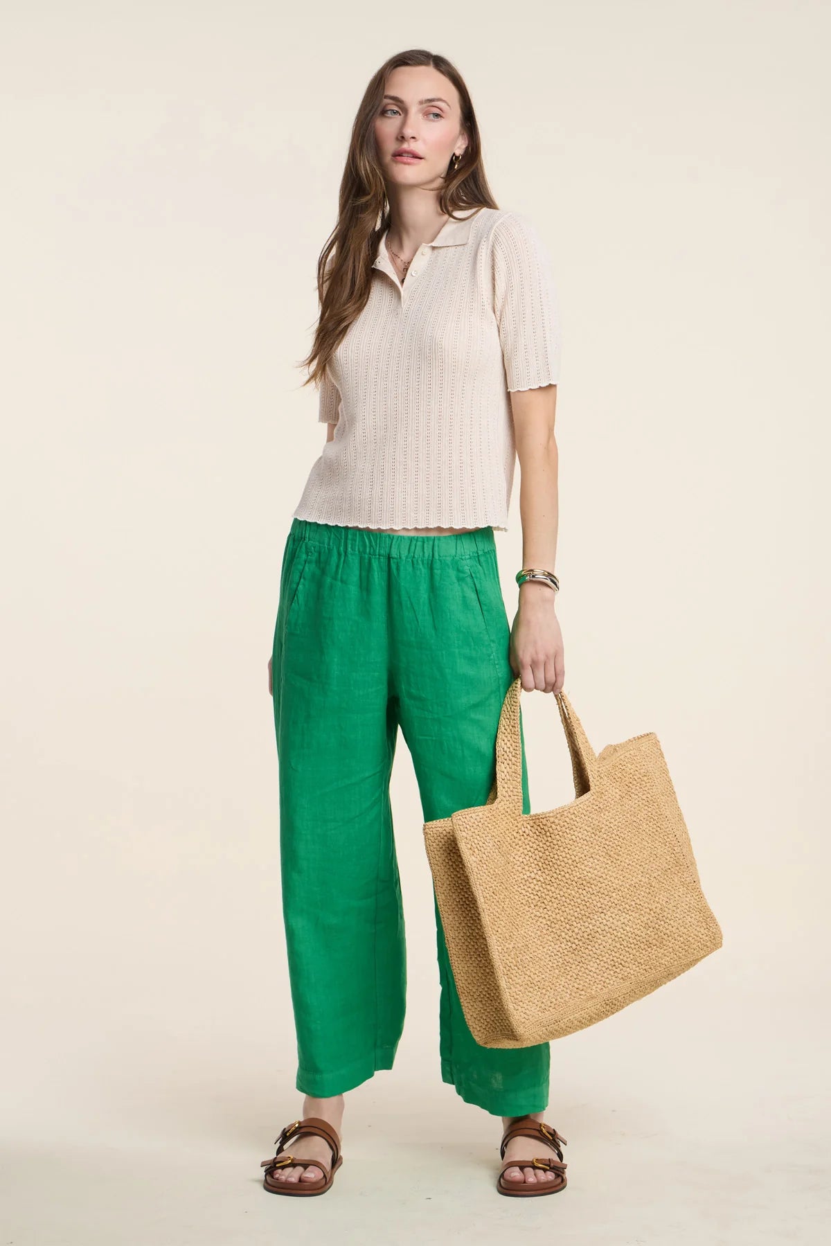 Woman holding a woven bag, wearing a beige shirt and green pants on a plain background