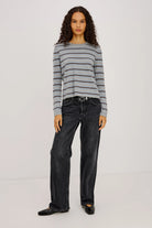 Woman wearing a gray and blue striped sweater with black jeans and a ballet flat on a plain background