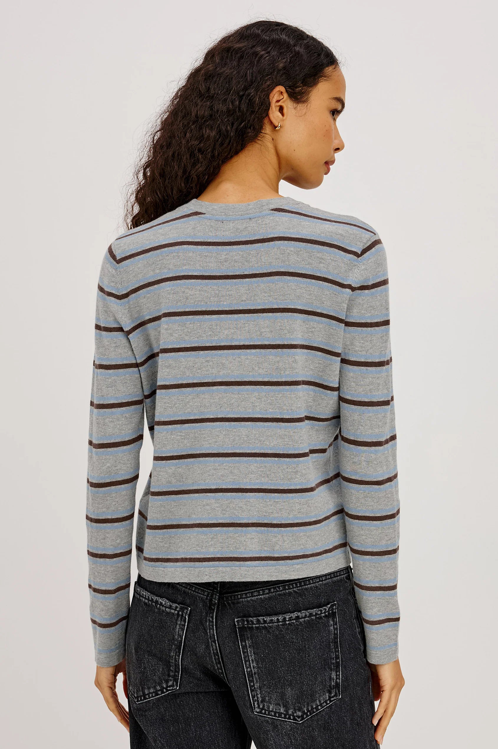 Woman wearing a gray and blue striped sweater with black jeans on a plain background from the back view.