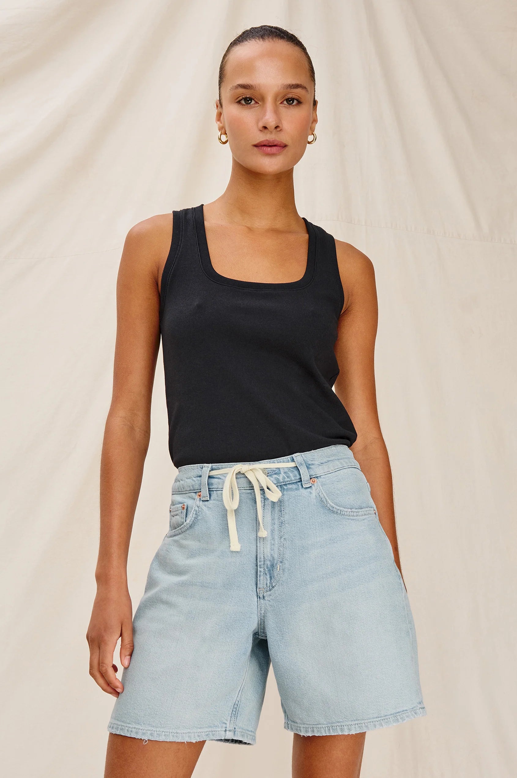 Woman wearing a black tank top and light blue denim shorts on a beige background