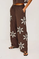 Brown wide-leg pants with white floral patterns worn by a person on a white background