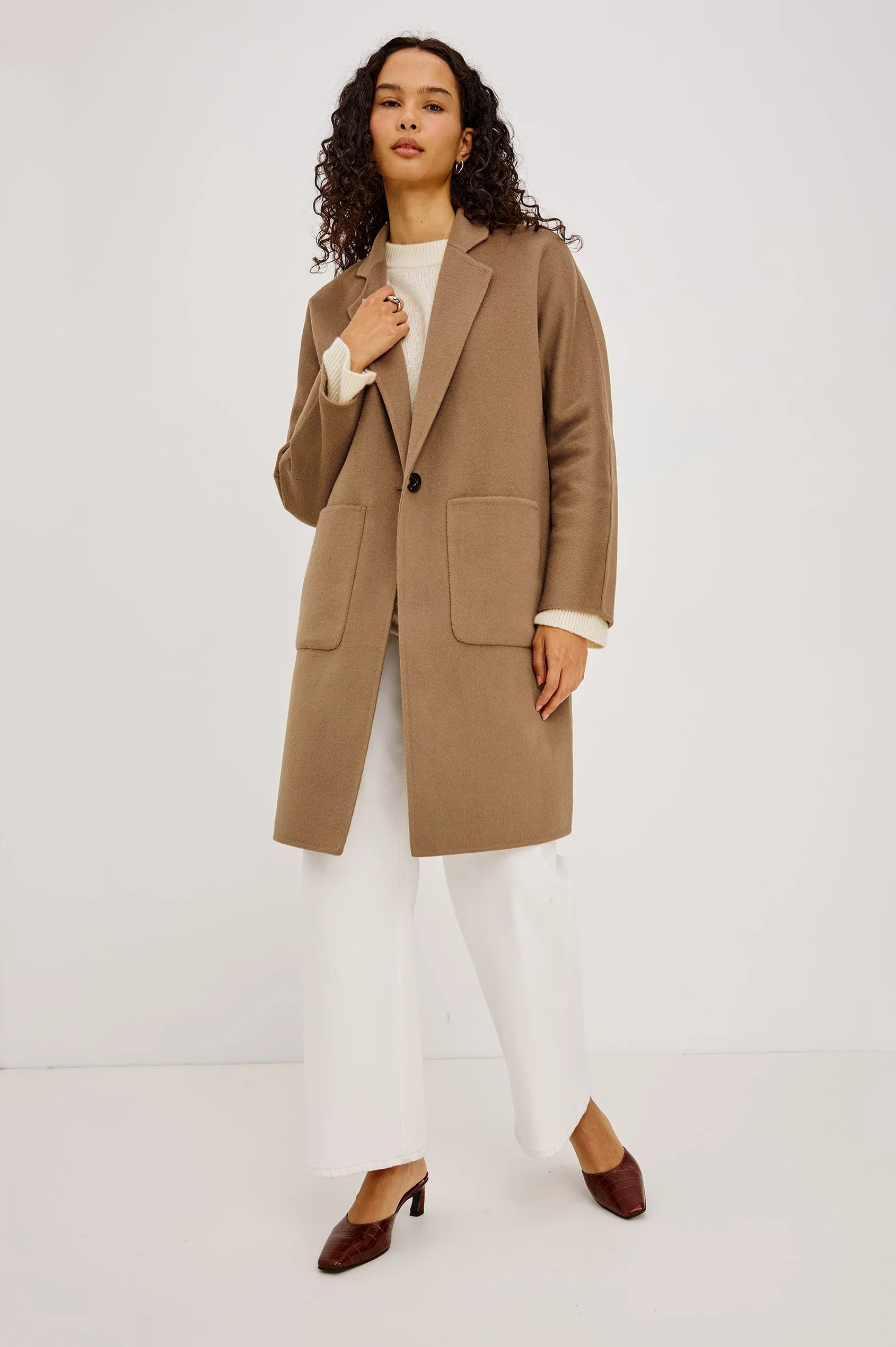 A woman modeling a camel brown wool-blend coat with a single-breasted button closure and large front patch pockets, worn over a cream top and white wide-leg trousers, against a plain light gray studio background.