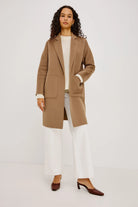A woman modeling a mid-length camel brown wool-blend coat over a cream top and white wide-leg trousers, paired with dark brown pointed-toe heeled mules, against a plain white studio background.