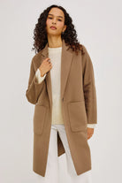 A woman with curly dark hair models a mid-length camel brown coat over a cream knit top and white pants, photographed against a plain light gray studio background.