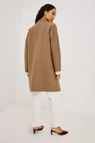 A woman wearing a camel brown wool-blend coat with white trousers and dark brown mule heels, photographed from the back in a minimalist white studio setting.