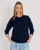 A woman modeling a fuzzy, long-sleeve crewneck sweater in dark navy blue, paired with light blue jeans, against a plain light gray background.