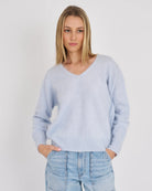 A woman wearing a light blue v-neck knit sweater and light-wash blue jeans, standing against a plain light gray studio background.
