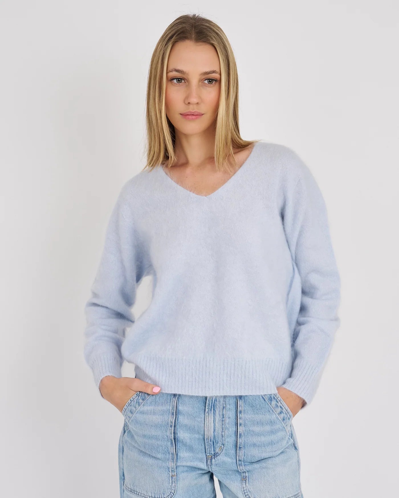 A woman wearing a light blue v-neck knit sweater and light-wash blue jeans, standing against a plain light gray studio background.