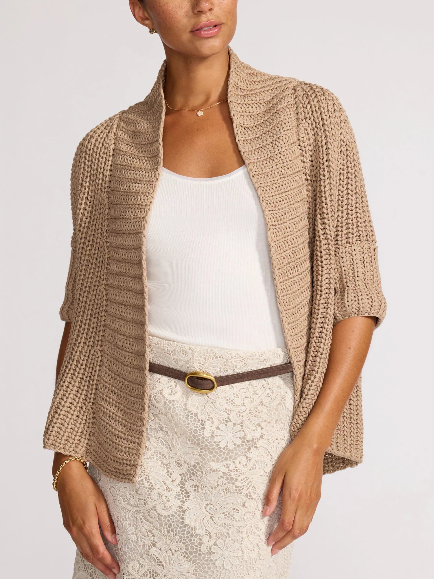 Woman wearing a beige knitted cardigan over a white top and skirt on a plain background