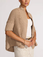 Woman wearing a beige knitted poncho over a white top on a plain background