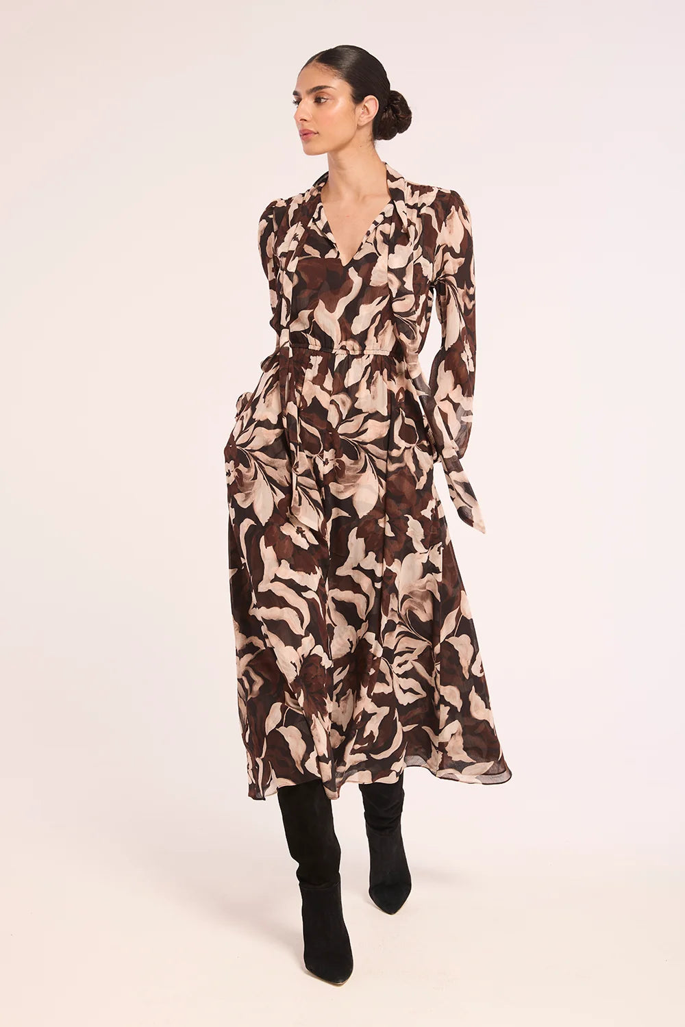 A woman modeling a long-sleeved midi dress with a brown, beige, and cream abstract print, paired with black knee-high boots, against a plain light background.