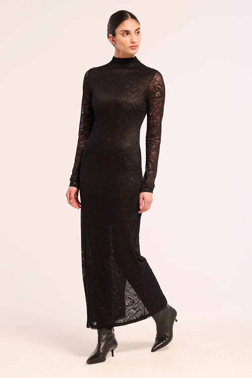 A woman modeling a black long-sleeve mock-neck lace maxi dress with black leather ankle boots, standing against a plain light beige studio background.