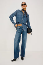 A model wearing a matching blue denim set consisting of a long-sleeve button-up shirt and wide-leg jeans, styled with a black neck scarf, sunglasses, and a small black handbag against a plain white studio background.