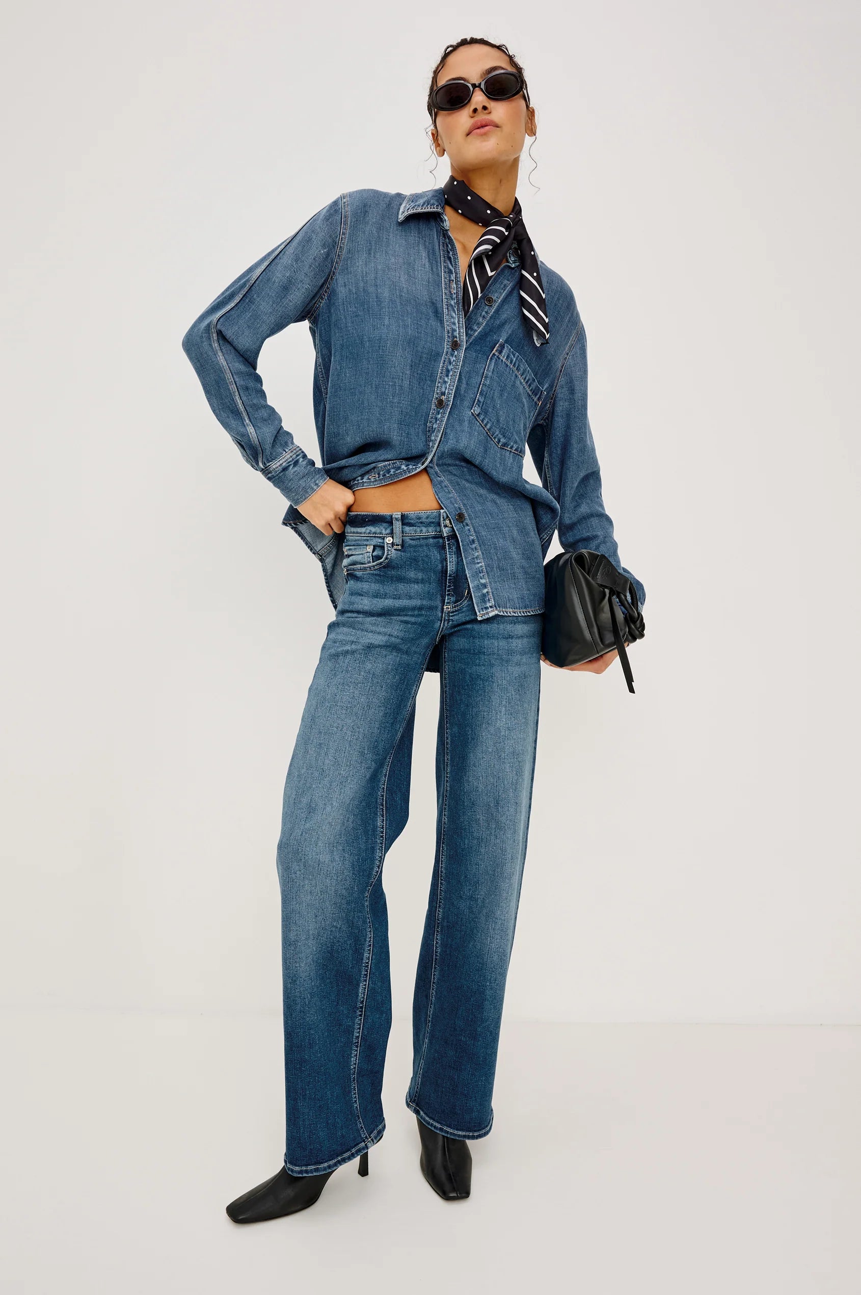 A model wearing a matching blue denim set consisting of a long-sleeve button-up shirt and wide-leg jeans, styled with a black neck scarf, sunglasses, and a small black handbag against a plain white studio background.